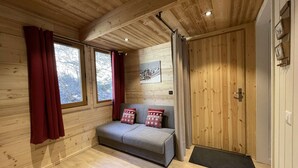 A cozy room with wooden walls, a grey sofa with patterned cushions, a window with red curtains, and a door with a wooden panel. - N°18 : 6/8 people flat incl. swimmingpool, sauna, hammam (CHAMPAGNY EN VANOISE)