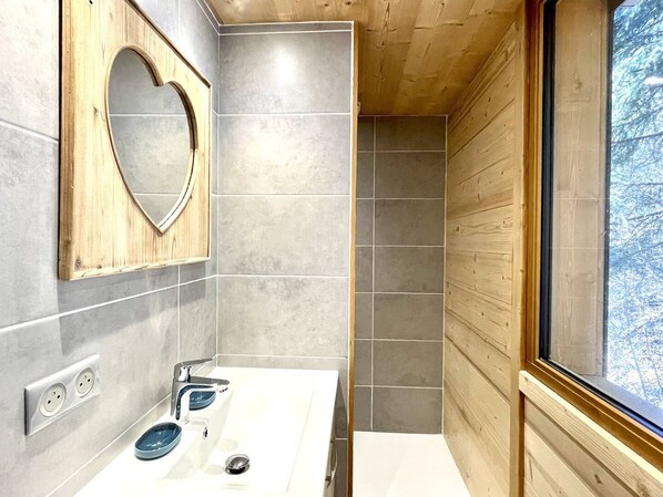 Un baño moderno con un espejo con forma de corazón, un lavabo doble con lavamanos azul y una ventana con vistas a árboles.