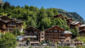 A cluster of wooden buildings with balconies, labeled "CLUB ALPINA," surrounded by lush greenery and mountains.