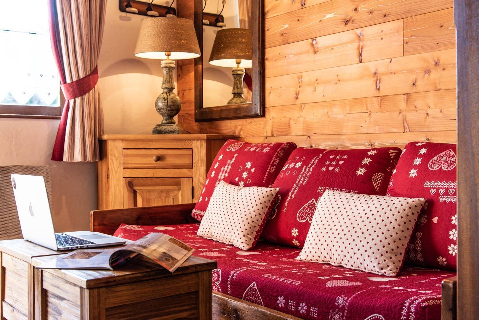 A cozy room with a wooden wall, a red sofa with patterned cushions, a wooden nightstand with a laptop, and a lamp.
