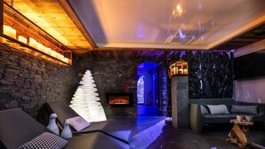 A modern spa room with a stone wall, a fireplace, a relaxation area with a massage table, and a seating area with a sofa and a decorative tree sculpture.