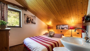 A cozy bedroom with a wooden ceiling, a large bed with striped bedding, a bedside lamp, a wall-mounted painting of bears, and a window with curtains.