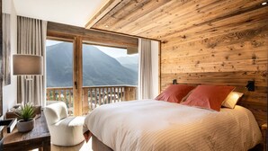 A bedroom with a large bed, wooden ceiling, a view of mountains, a chair, and a table with a plant.