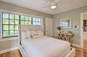 A bedroom with a large bed, a ceiling fan, a window with blinds, a bedside table with a mirror, and a door.