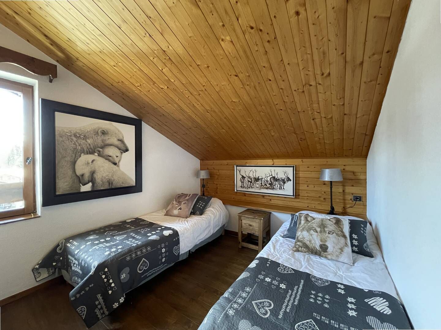 Una habitación acogedora con techo inclinado, dos camas con ropa de cama estampada, un cuadro con animales enmarco y una lámpara sobre una mesita de noche de madera.