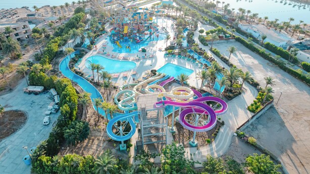 Vista aérea de un parque acuático con múltiples toboganes y piscinas, rodeado de palmeras y zonas de playa.