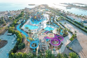 Vista aérea de un parque acuático con múltiples toboganes y piscinas, rodeado de palmeras y zonas de playa.