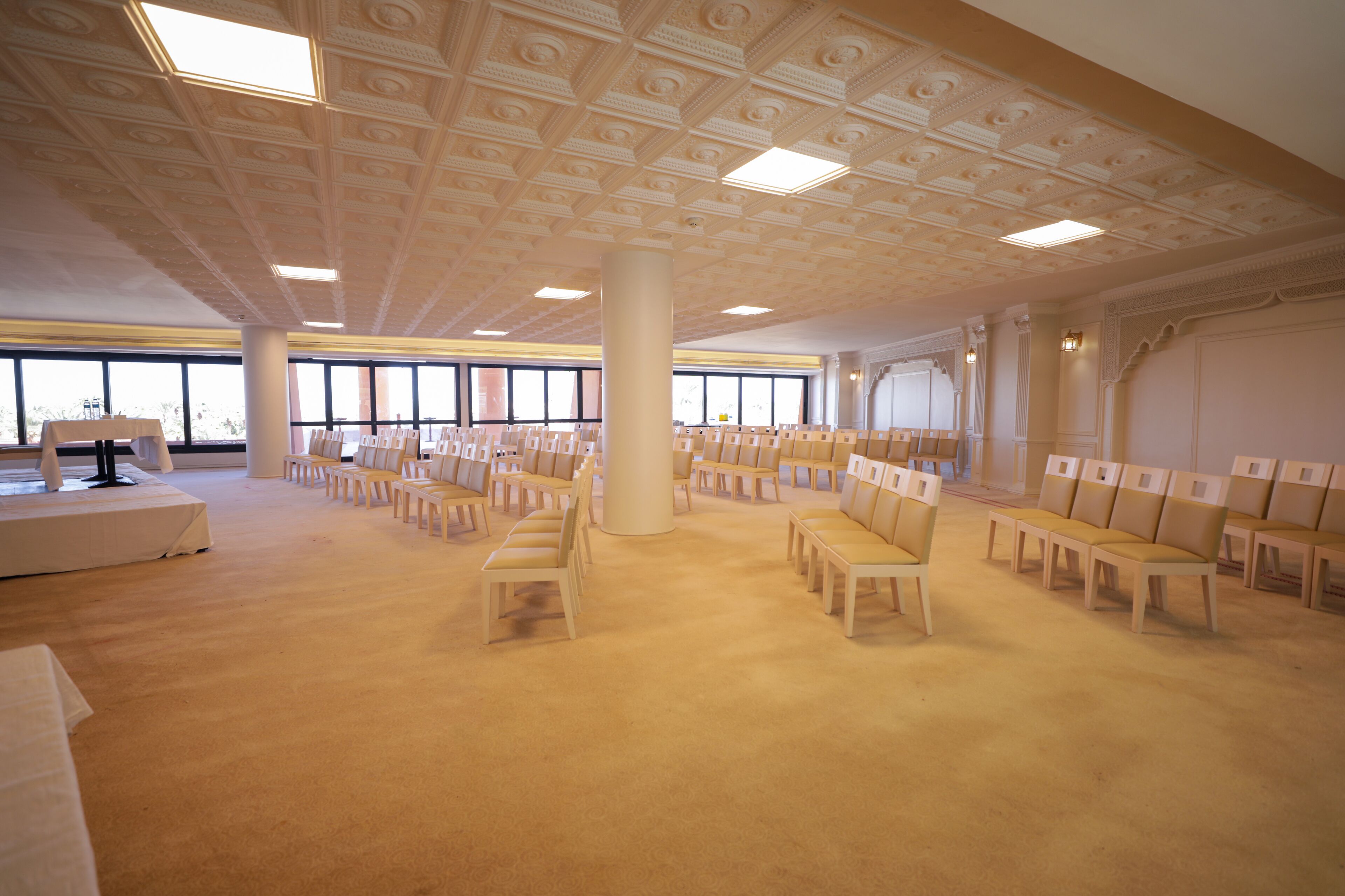 Una habitación amplia con filas de sillas de madera, un escenario con atril y ventanas grandes que dejan entrar luz natural.