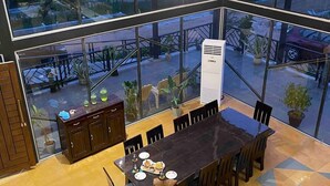 A dining area with a large table and chairs, a wooden cabinet, and a view of the outdoors through large windows. - Bliss Farm Stay-JASMINE (Amangal)