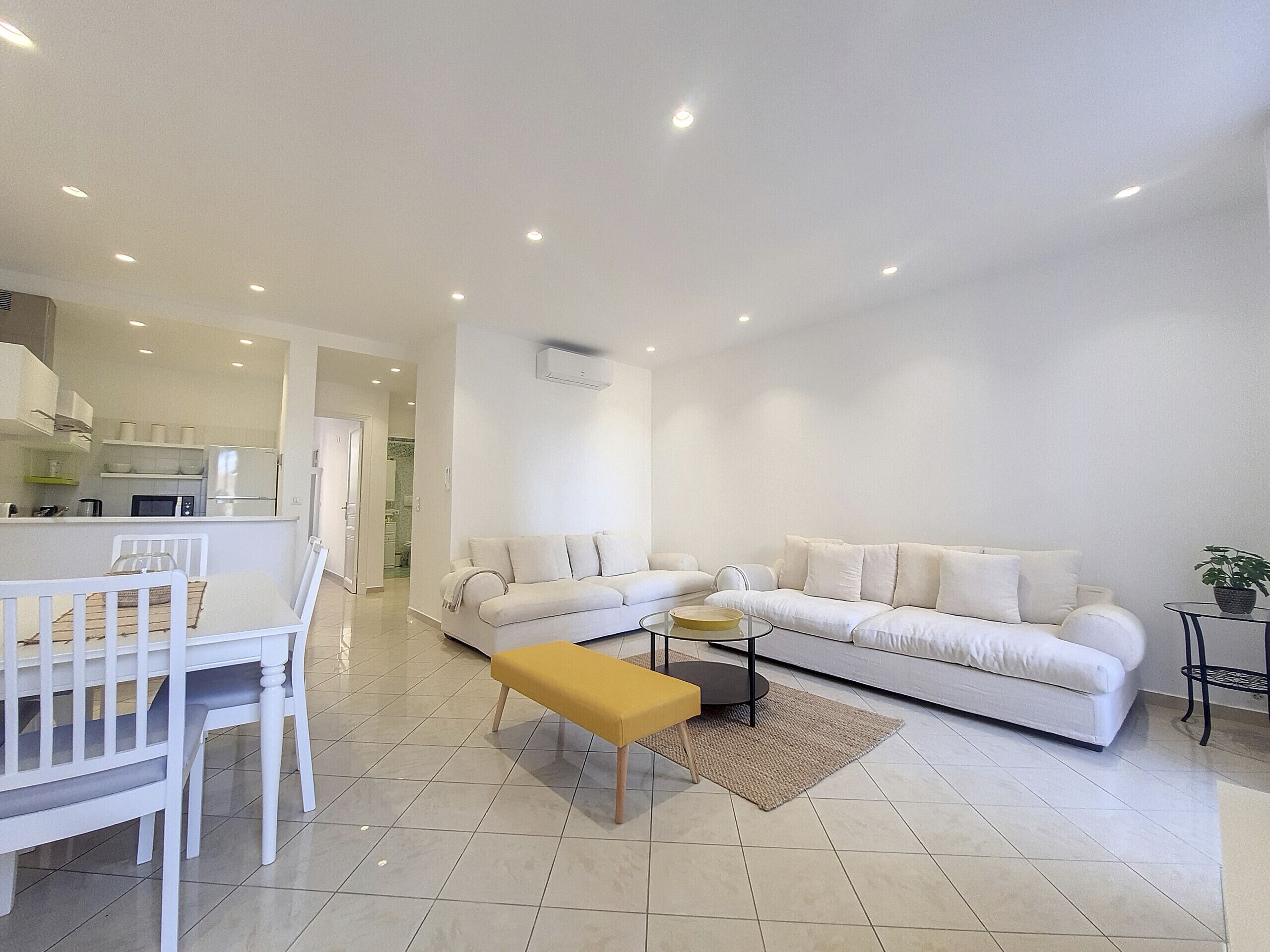 A modern living room with a white sofa, a dining table with chairs, and a kitchen area in the background.