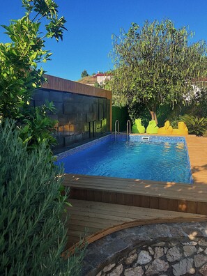 Una piscina rectangular con borde de azulejos, rodeada por una terraza de madera y un muro de piedra.