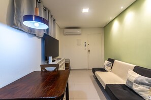 A living room with a wooden dining table, a sofa with cushions, a television, and an air conditioning unit. - ana cristina (Maceió)