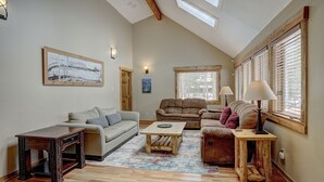 A spacious living room with a high ceiling, wooden beams, a large window with blinds, a sofa, a coffee table, and a fireplace. - Ziggy Bear Lodge (Breckenridge)