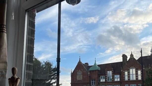 Ein Fenster mit Blick auf ein Gebäude, eine Vase mit Blumen und eine Fernbedienung auf einem Fensterbrett.