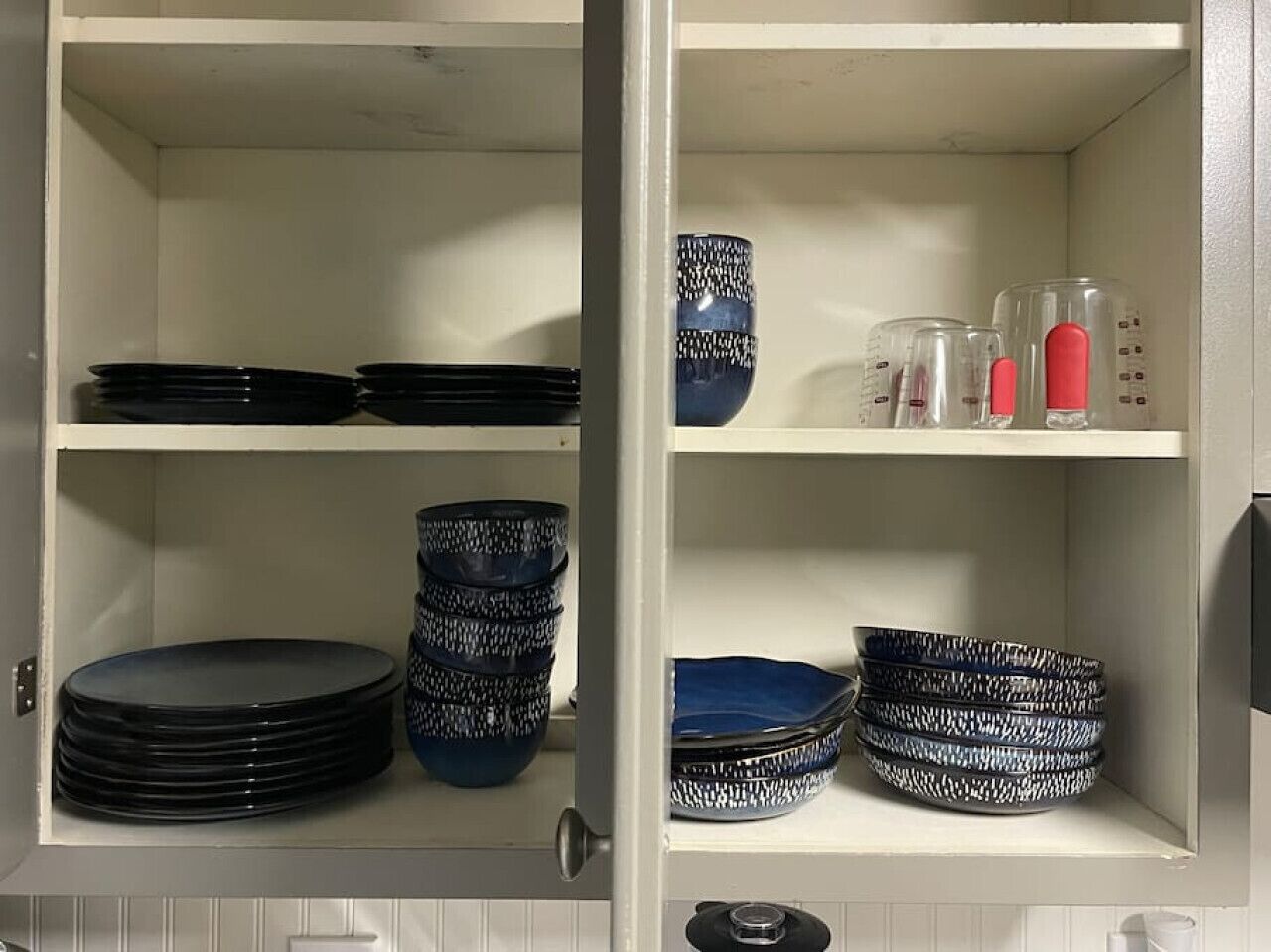 A kitchen cabinet with shelves holding black plates, blue and white patterned bowls, and clear measuring cups with red lids.