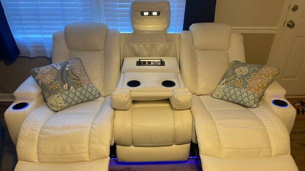 A reclining massage chair with a control panel and two footrests, placed in a room with a wooden floor and a window with blinds.