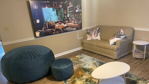 A living room with a large bean bag, a small ottoman, a round coffee table, and a sofa. A large framed picture hangs on the wall.