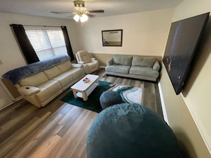 Una sala de estar con un sofá color beige, un sillón de tonos claros, una mesa de centro blanca y un asiento de bola verde. Hay un ventilador de techo, una ventana con persianas y un cuadro en la pared.