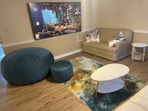 Una sala de estar con un televisor de pantalla plana grande, un sofá color beige, dos pufs oscuros, una mesita blanca y una alfombra colorida.