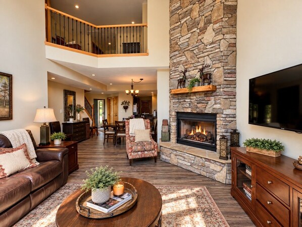 A spacious living room with a fireplace, a high ceiling, and a staircase leading to an upper level. - Seven Springs Condo Near Shuttle, Gas Fireplace (Champion)