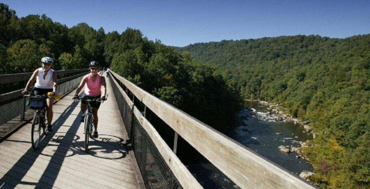 Deux cyclistes sur un pont en bois, avec une rivière et des collines boisées en arrière-plan.