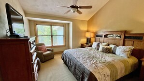 A bedroom with a large bed, a desk with a computer, a chair, a ceiling fan, and a window with blinds.