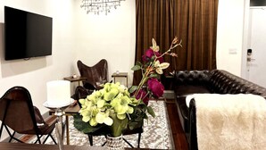 A modern living room with a flat-screen TV, a leather sofa, a dining table with a vase of flowers, and a patterned rug.