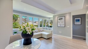 A modern living room with a glass table, white sofa, and large windows offering a view of the outdoors.