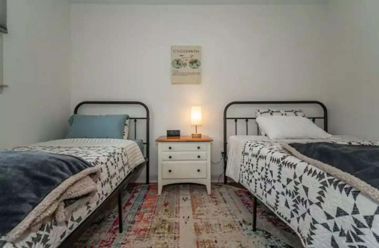 A small, tidy bedroom with two single beds, a nightstand with a lamp, and a wall-mounted sign.