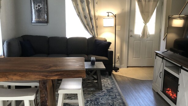 A cozy living area with a wooden dining table, white bar stools, a black sofa, a patterned rug, a fireplace, and a framed picture on the wall. - Sweet Escape Cottage on River  (Barton)