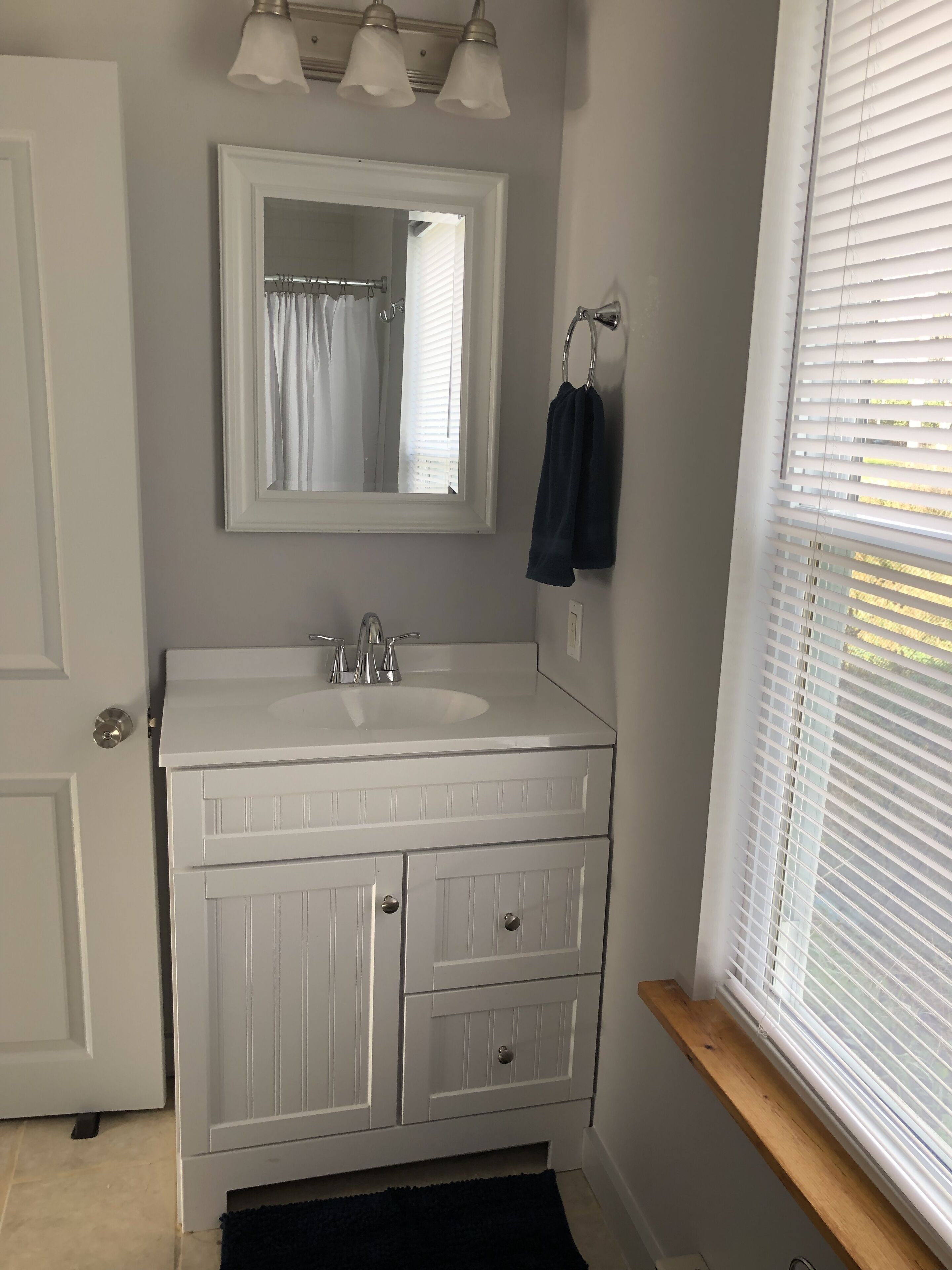 Une salle de bain avec un meuble-lavabo blanc, un miroir, un évier et une fenêtre avec des stores.