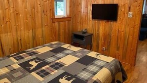 Une pièce aux murs en bois, un lit recouvert d’une couverture à carreaux, une petite table avec une lampe et un téléviseur fixé au mur.
