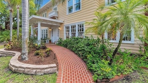 A two-story building with a covered porch, a brick pathway, and lush greenery including palm trees. - Walnut Room at ONE Inn, Comfy, Steps to Downtown St. Pete: Ideal for all guests! (Saint Petersburg)