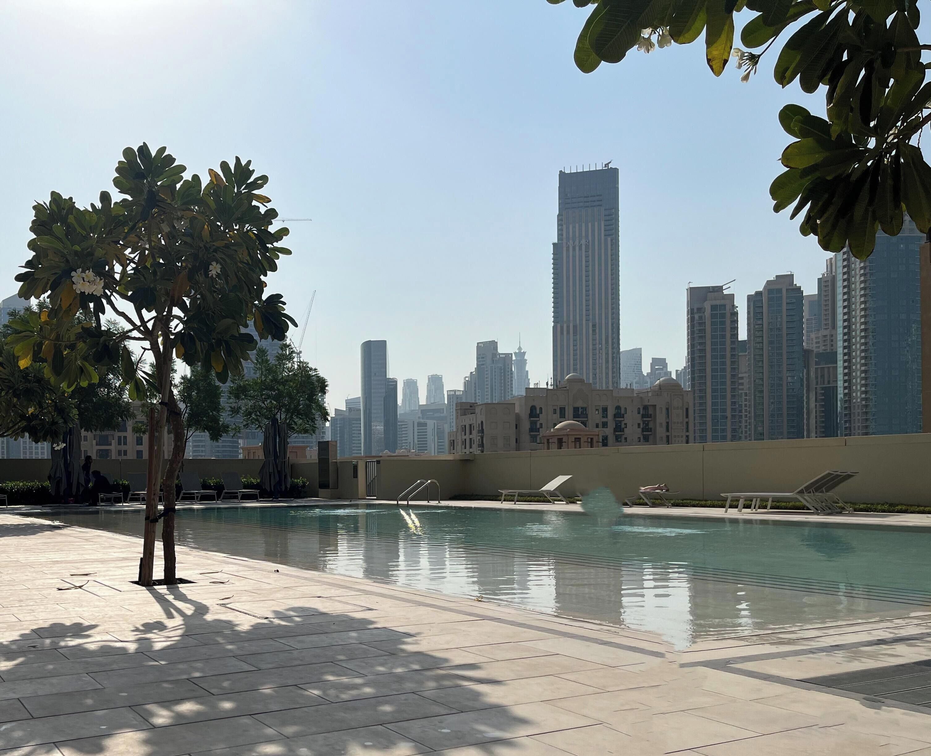 A swimming pool with lounge chairs, surrounded by trees and a cityscape in the background.