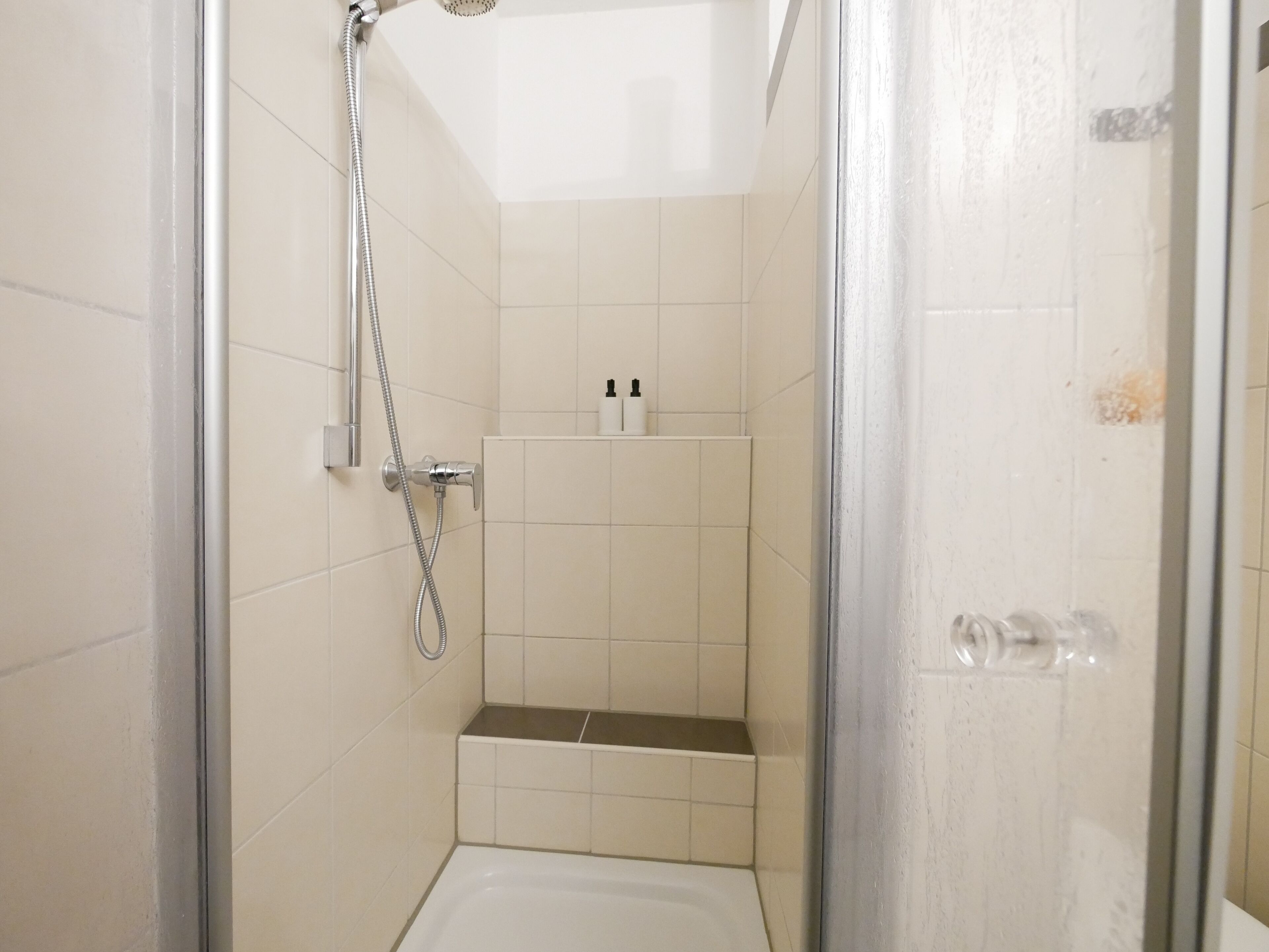 Een kleine, afgesloten douchecabine met douchekop en slang, betegelde wanden en een klein plankje met toiletartikelen.