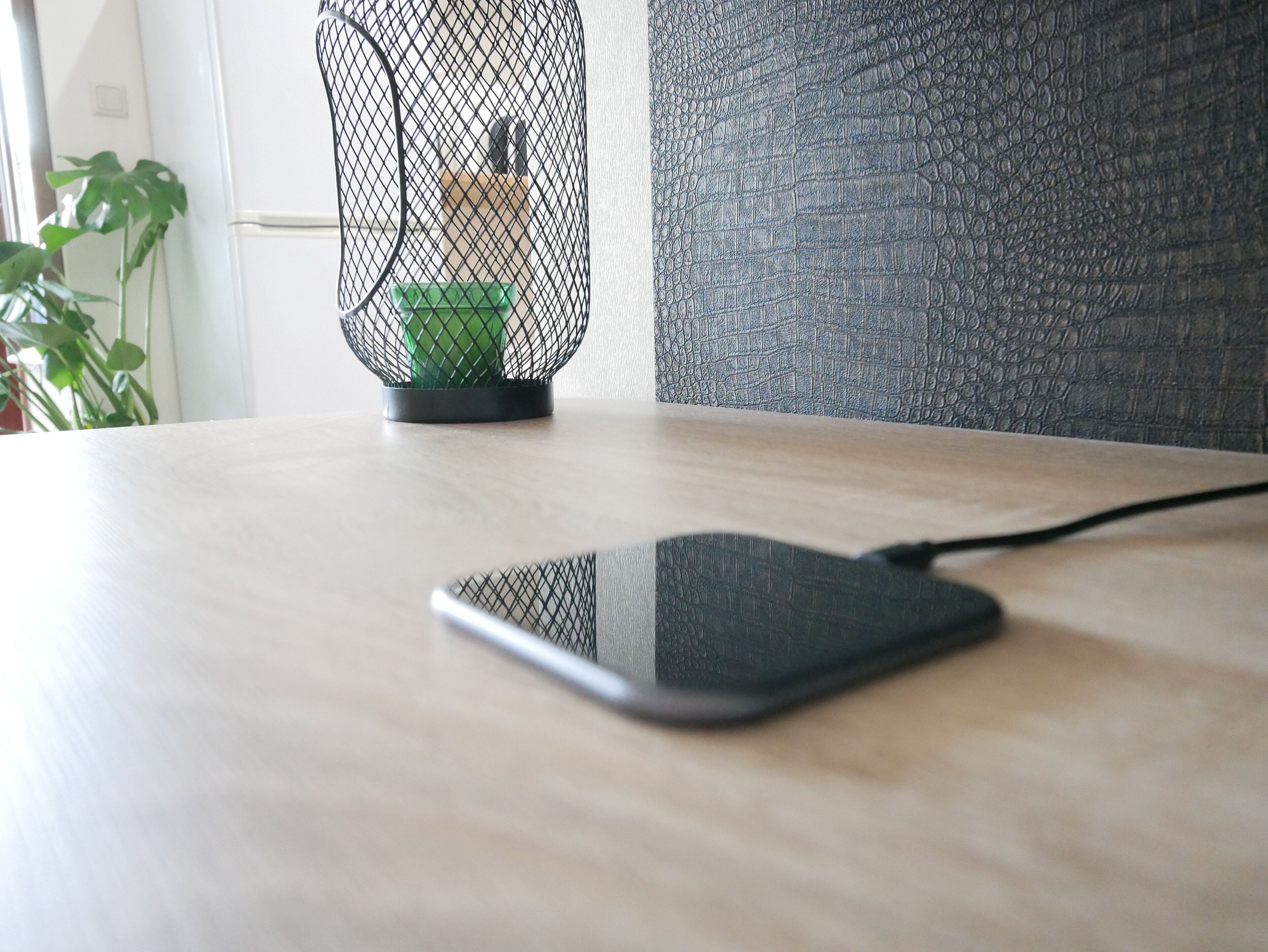 A smartphone on a wooden surface with a textured wall and a plant in the background.