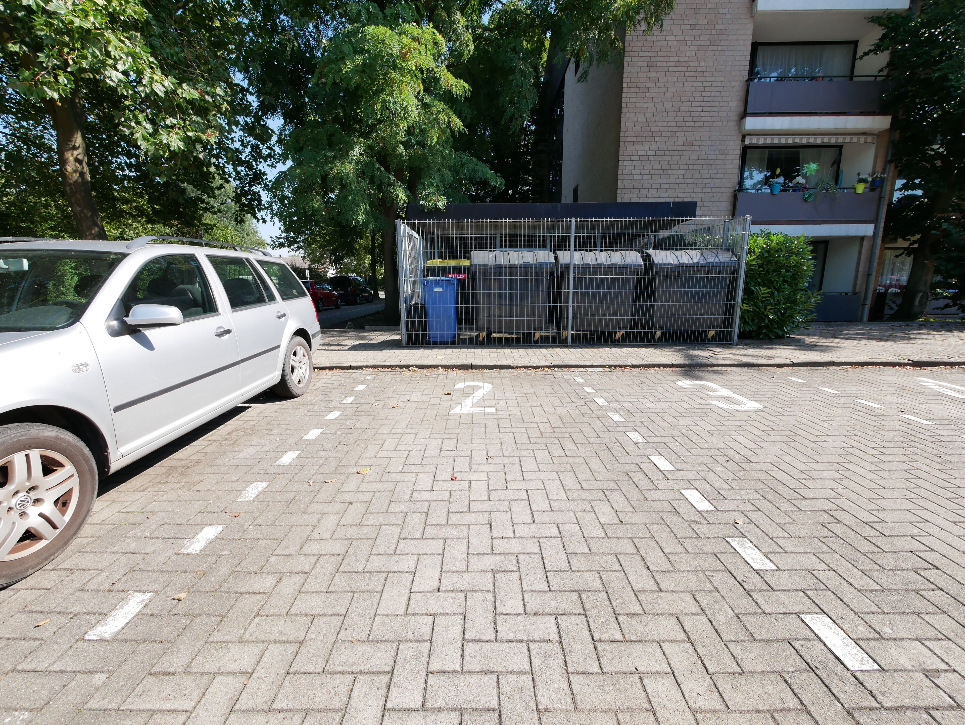 A parking area with a white car parked on the left. There is a fenced area with bins and a building with balconies in the background.