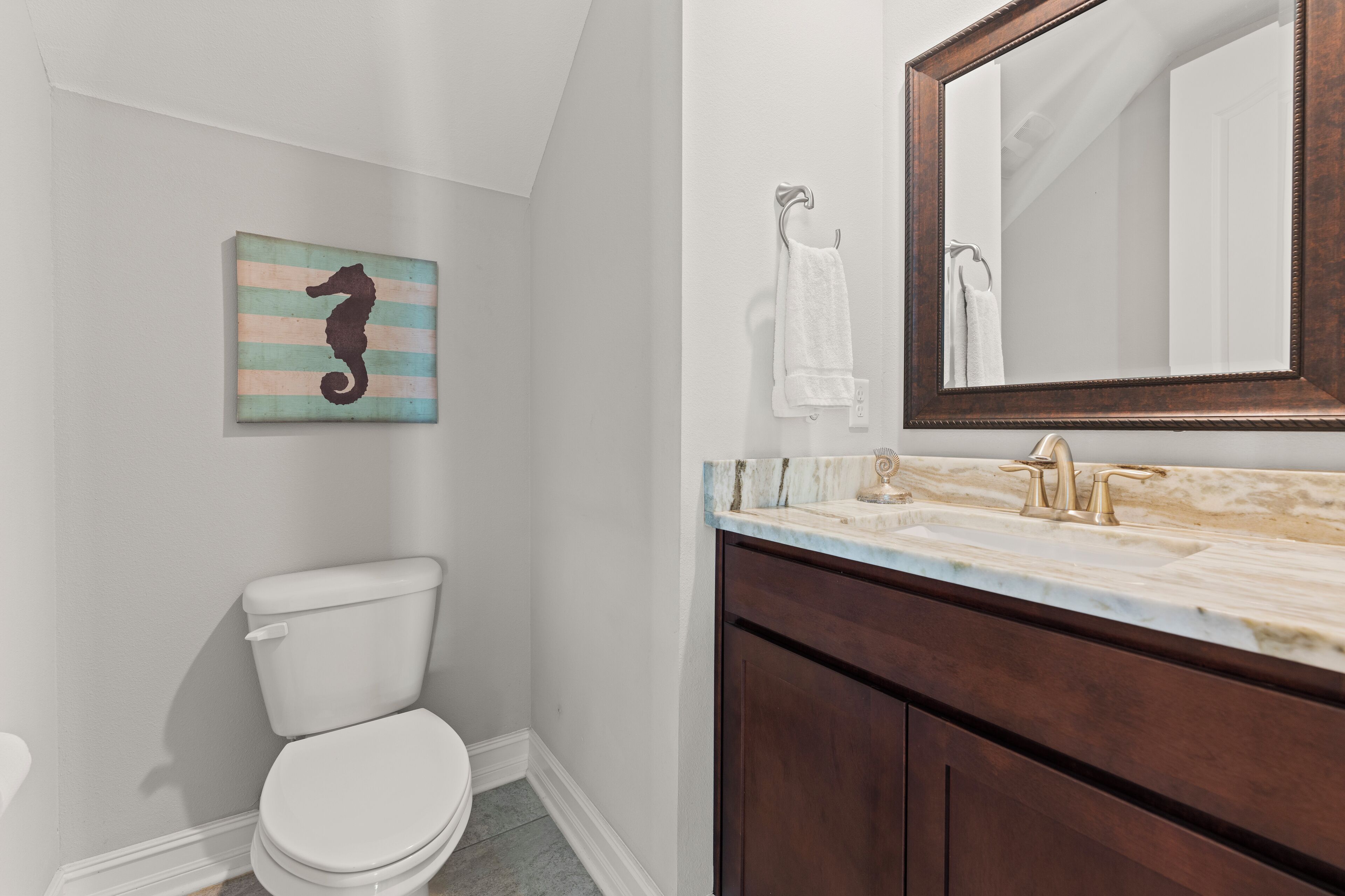 Un baño con inodoro blanco, tocador de madera con encimera de mármol, espejo y una obra de arte con motivos de caballito de mar en la pared.