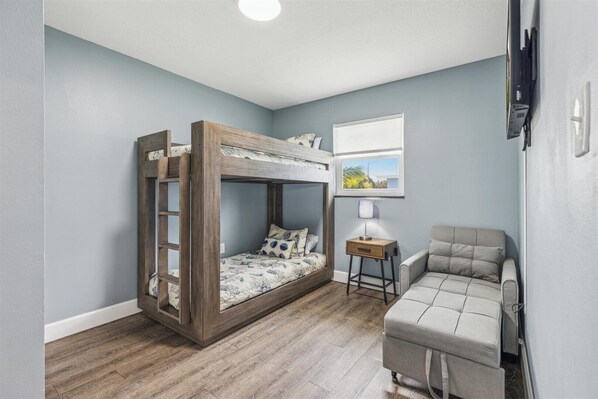 A bedroom with a bunk bed, a bedside table with a lamp, a sofa, and a window with a view of greenery.