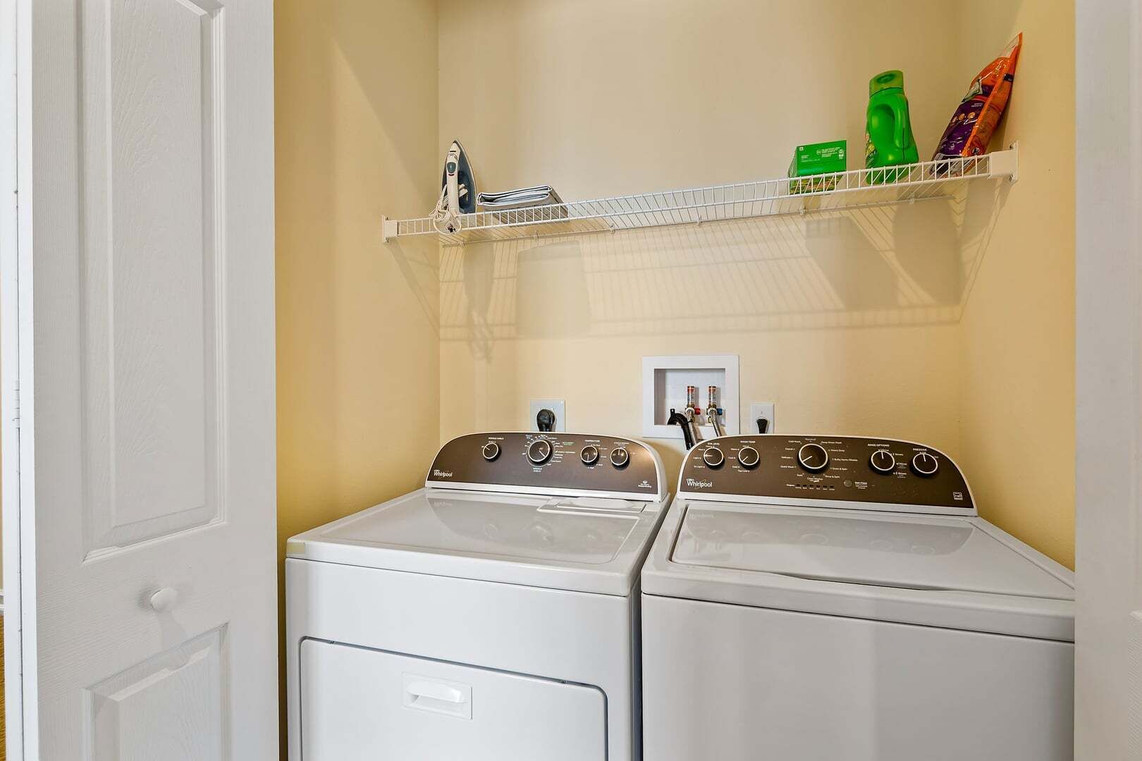 A laundry room with two Whirlpool washers and dryers, a shelf with cleaning supplies, and a door to the left.