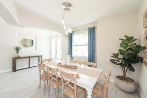 Uma sala de jantar com mesa branca, cadeiras de madeira, luminária pendente e planta em vaso.