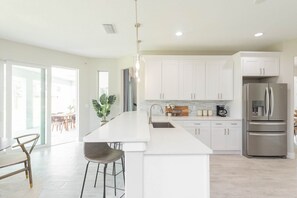 A modern kitchen with white cabinetry, a stainless steel refrigerator, and a bar area with stools. - 313 Surf Drive (Cape Canaveral)