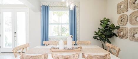 A dining room with a white table, wooden chairs, a chandelier, and a large window with blue curtains.