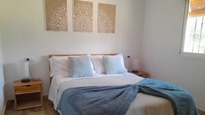 Un dormitorio con una cama, dos mesitas de noche, una ventana con persianas y tres obras de arte en la pared.
