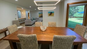Un comedor moderno con una mesa de madera y sillas tejidas, una sala de estar al fondo y un ventanal con vistas a zonas verdes.