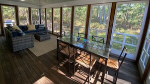 Un solárium con una mesa y sillas de vidrio, un sofá y ventanas amplias que dan a un bosque.