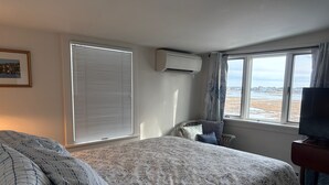A hotel room with a large bed, a television, a window with a view of the beach, and an air conditioning unit.