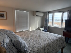 A hotel room with a large bed, a television, a window with a view of the beach, and an air conditioning unit.