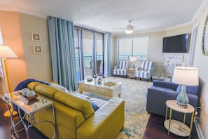 A living room with a yellow sofa, a glass coffee table, a blue armchair, a striped armchair, a television, and a balcony with curtains.