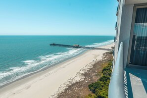 Una playa con un muelle que se adentra en el océano, una orilla arenosa y un edificio con balcón con vistas a la escena.
